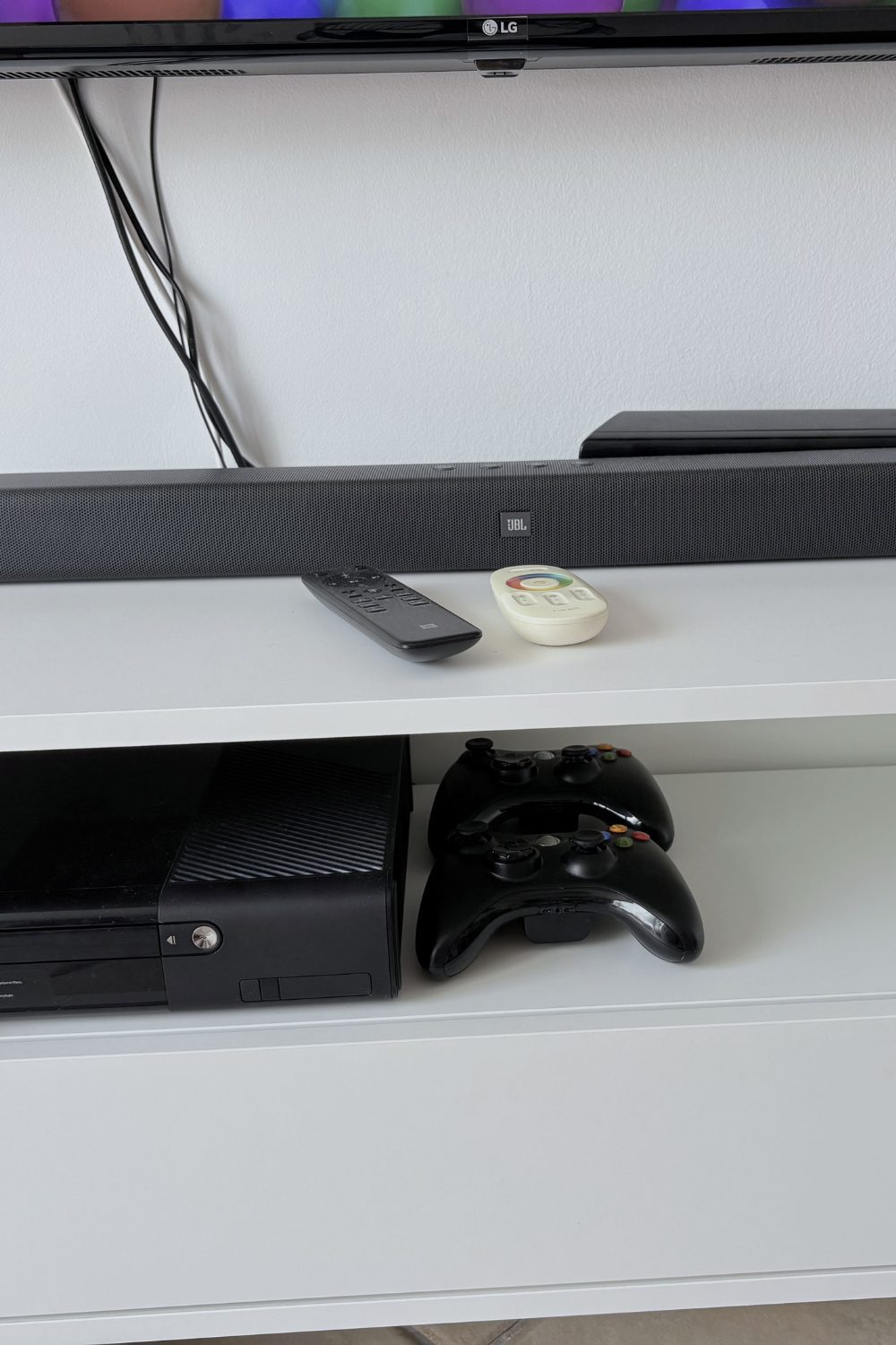 A white TV console with a soundbar, remotes, a games console and cables running down the wall