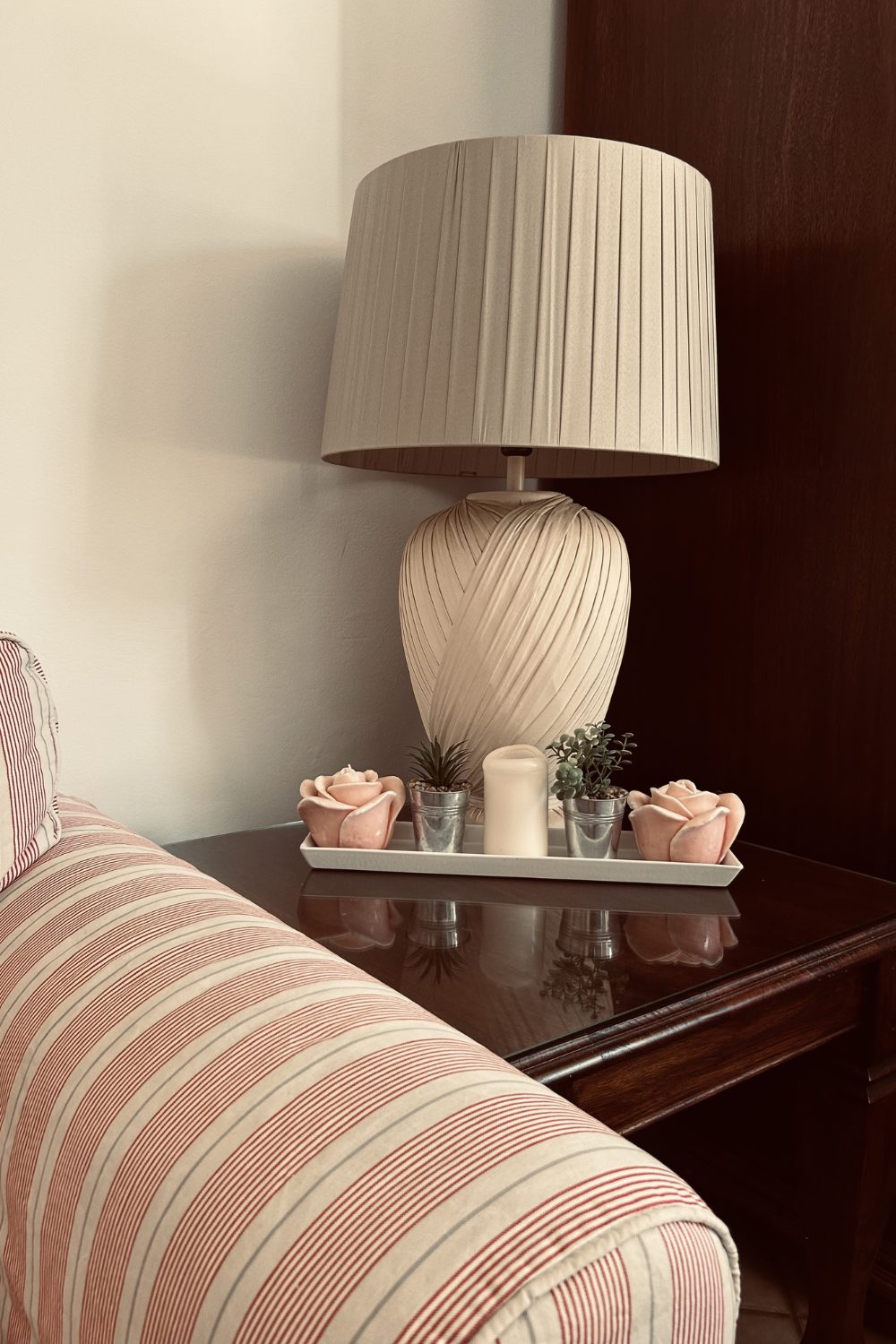A pleated cream table lamp on a dark wood side table beside a striped armchair
